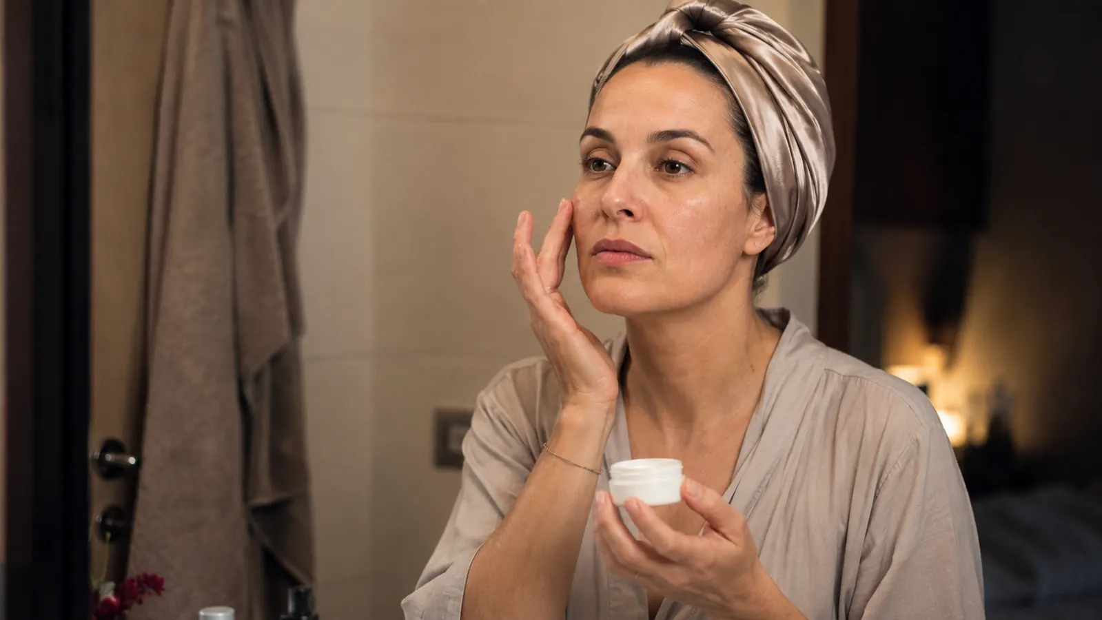 Woman with glowing skin in a soft-lit bathroom applying overnight mask from a tub before bed, wearing a hair wrap