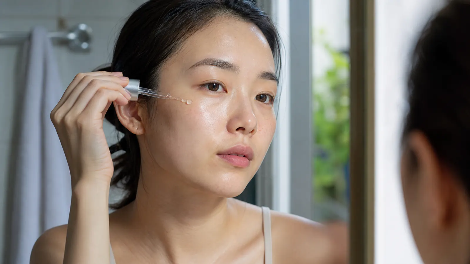 Woman with glowing, hydrated skin applying serum in a bright bathroom mirror, K-beauty morning routine